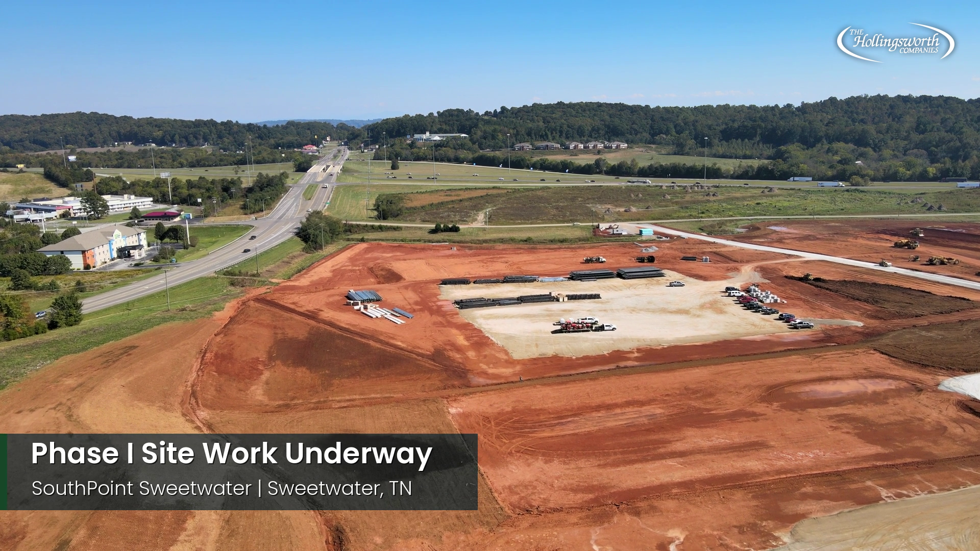 Drone view of graded red clay building pads and a gravel pad with construction materials at SouthPoint Tennessee in Sweetwater, with Interstate 75 and a hotel to the left and wooded hills beyond. The Hollingsworth logo appears in the corner.